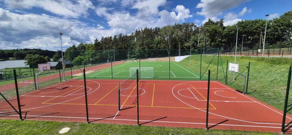 Ogrodzone boiska sportowe z czerwoną nawierzchnią do koszykówki i siatkówki oraz zielonym boiskiem piłkarskim. Obiekt otaczają trawiaste zbocza, drzewa i jezioro.